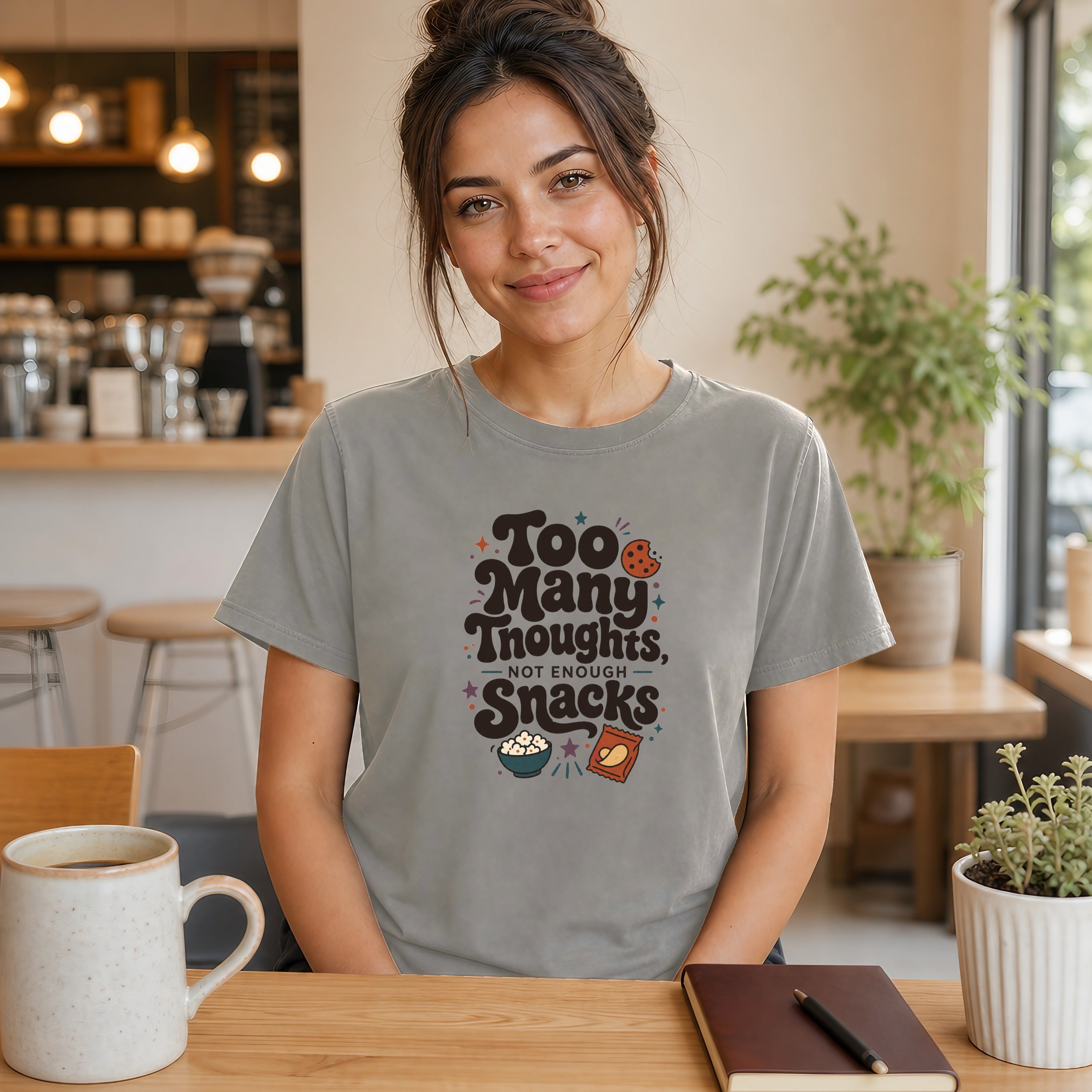 Woman wearing a gray t-shirt with "Too Many Thoughts, Not Enough Snacks" text and snack graphics.