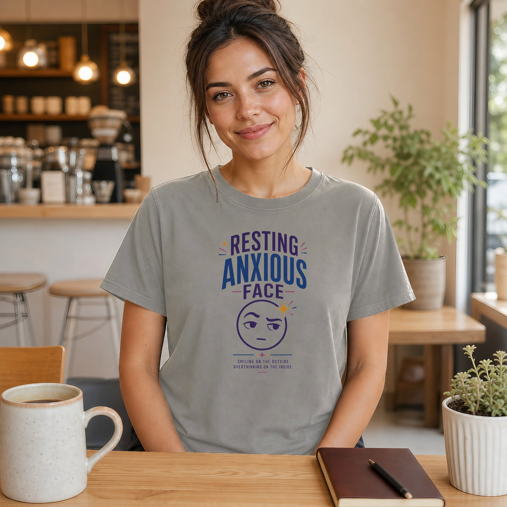 Woman in gray t-shirt with "Resting Anxious Face" graphic at cafe table.