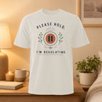 White t-shirt with "PLEASE HOLD, I'M REGULATING" text and pause symbol.