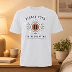 White t-shirt with "PLEASE HOLD I'M REGULATING" text and pause symbol.