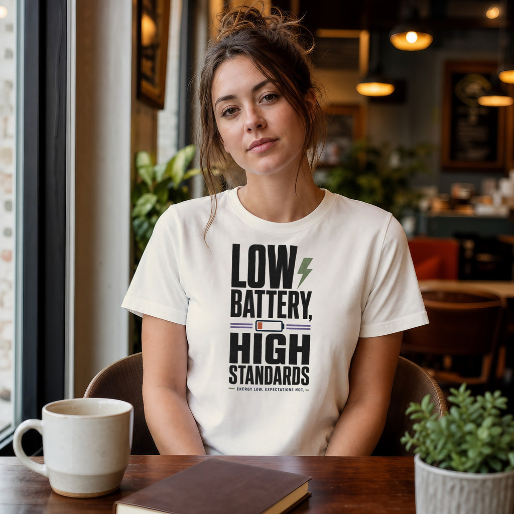Woman wearing cream t-shirt with "Low Battery, High Standards" text.